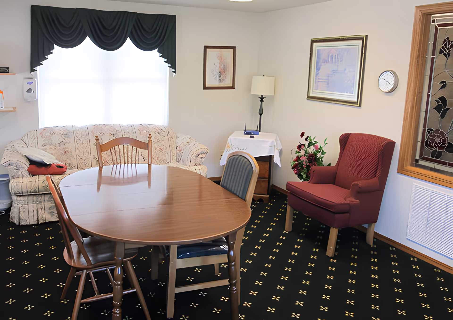 A cozy living room with a wooden dining table surrounded by four chairs, a floral patterned sofa under a window with dark green valance curtains, a red armchair, a small side table with a lamp and a router, framed artwork on the walls, and a decorative stained glass window on the right.