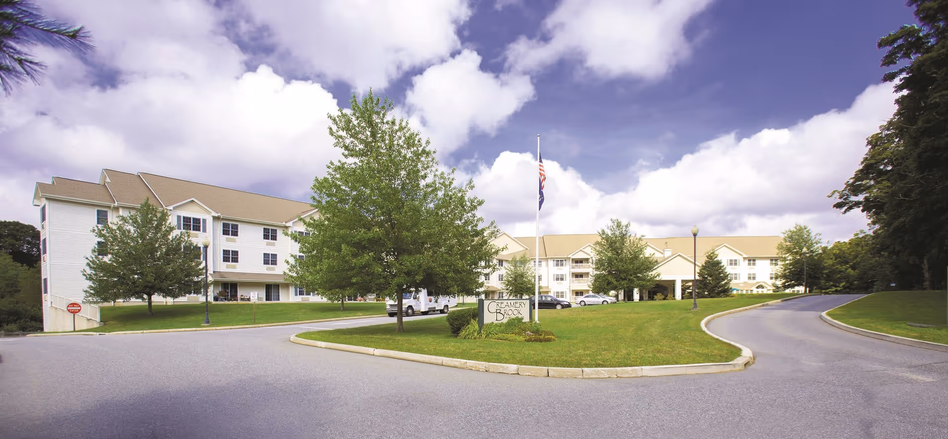 Exterior view of Creamery Brook Village, a multi-story senior living facility with a beige roof and white siding, surrounded by green lawns and trees under a partly cloudy sky. A driveway curves around a grassy area with a sign that reads 'Creamery Brook'.