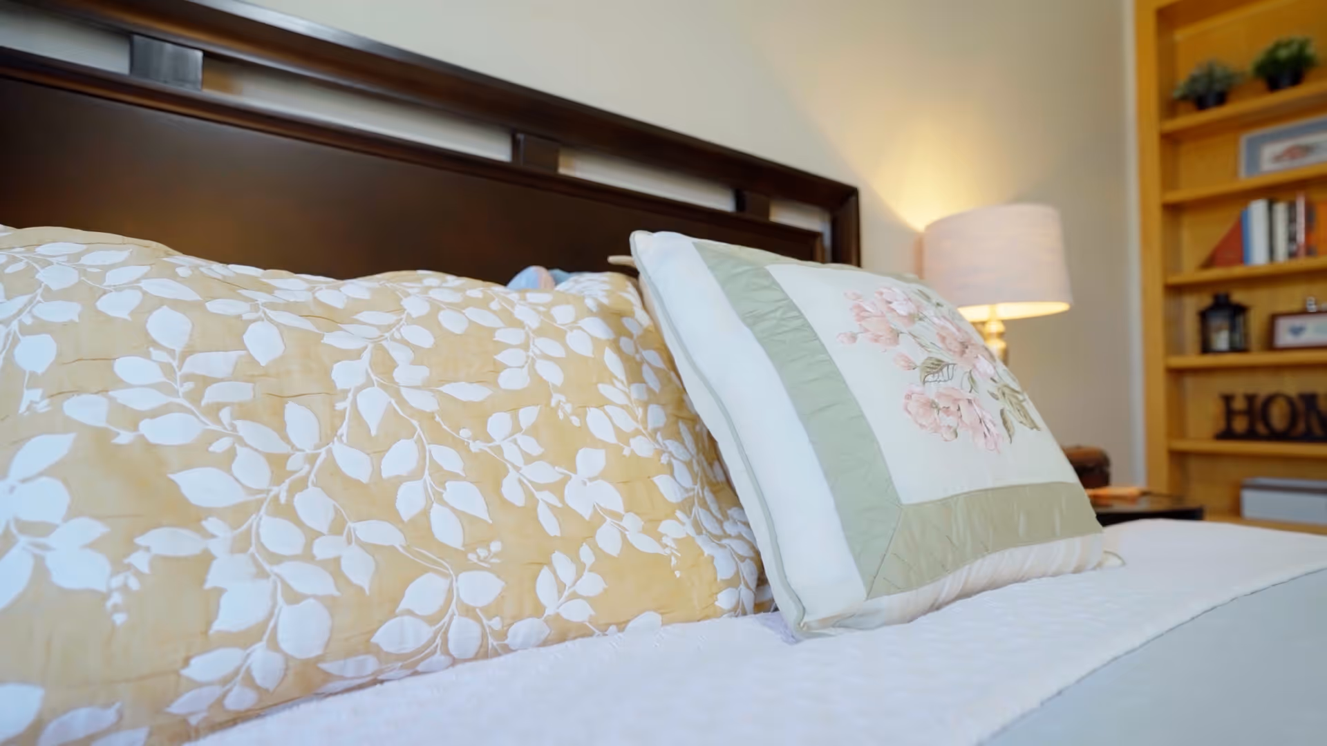Close-up view of a bed with decorative pillows, including one with a floral pattern and another with a white leaf design on a beige background. A wooden headboard is visible behind the pillows, and a lit lamp and bookshelf with decor items are in the background.