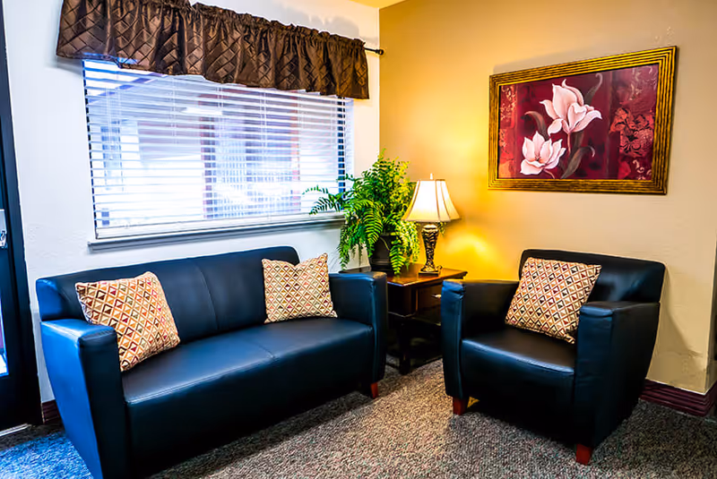 A cozy seating area with a black leather loveseat and matching armchair, each adorned with patterned throw pillows. Between them is a wooden side table holding a decorative lamp and a green potted plant. A window with blinds and a dark valance is behind the loveseat, and a framed floral painting hangs on the beige wall.