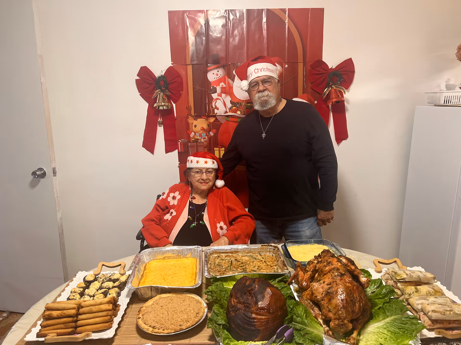 An elderly woman and man wearing Santa hats stand behind a table filled with various holiday dishes including a roasted turkey, ham, pie, and other side dishes. The woman is seated and wearing a red cardigan with floral designs, while the man stands beside her with his hand on her shoulder. The background features Christmas decorations including red bows and a festive poster with holiday characters.
