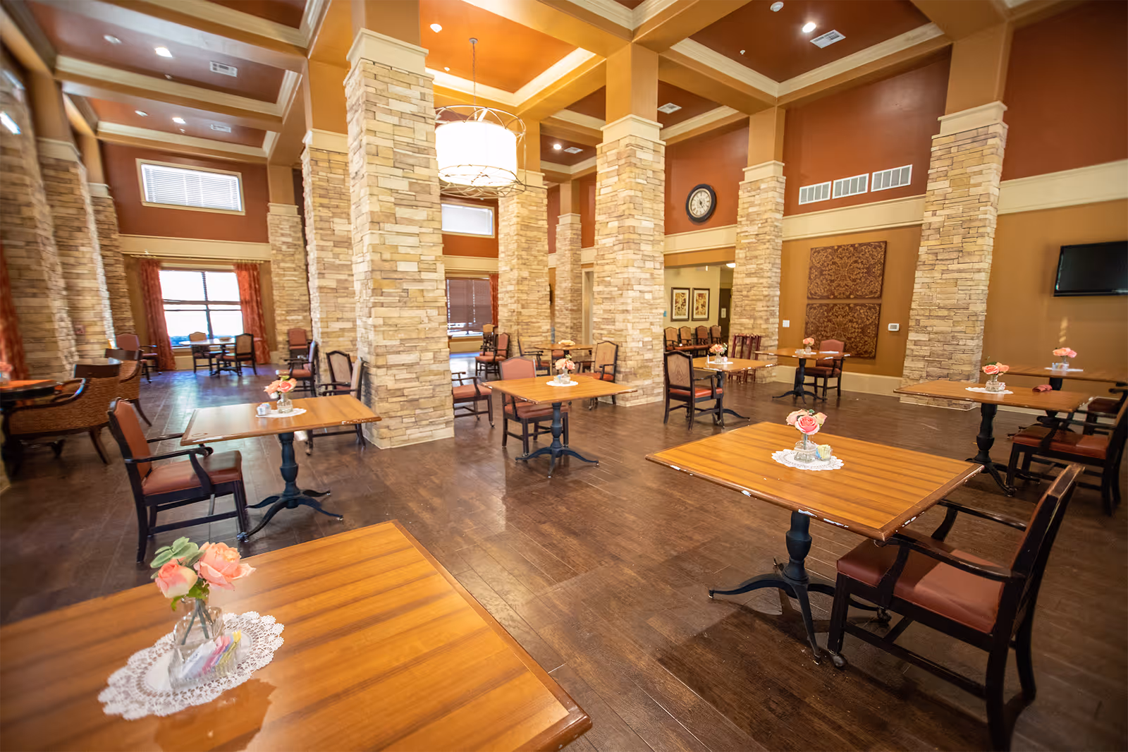 Spacious dining room with wooden tables and chairs arranged neatly. Each table has a small vase with pink flowers on a doily. The room features tall stone pillars, warm brown and beige walls, a large hanging light fixture, and a clock on the wall. Large windows with red curtains allow natural light to enter the room.