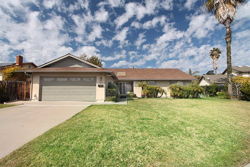 Single-story suburban ranch-style house with a two-car garage and a large front lawn under a partly cloudy sky.
