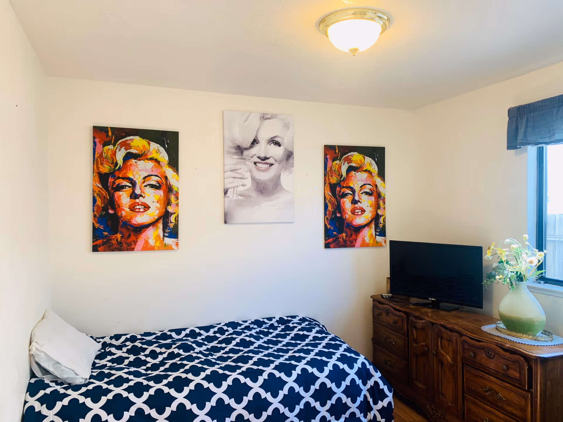 A bedroom with a single bed covered in a blue and white patterned bedspread. Above the bed are three wall art pieces featuring Marilyn Monroe, two colorful portraits on the sides and one black and white portrait in the center. To the right is a wooden dresser with a flat-screen TV and a vase with flowers. A window with a blue valance is visible on the right wall.