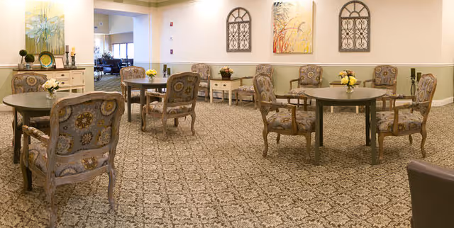 A spacious room with patterned carpet and several round tables surrounded by upholstered chairs with floral designs. The walls are decorated with framed artwork and decorative window-like wall hangings. There is a sideboard with decorative items and flowers, and the room has a warm, inviting atmosphere.