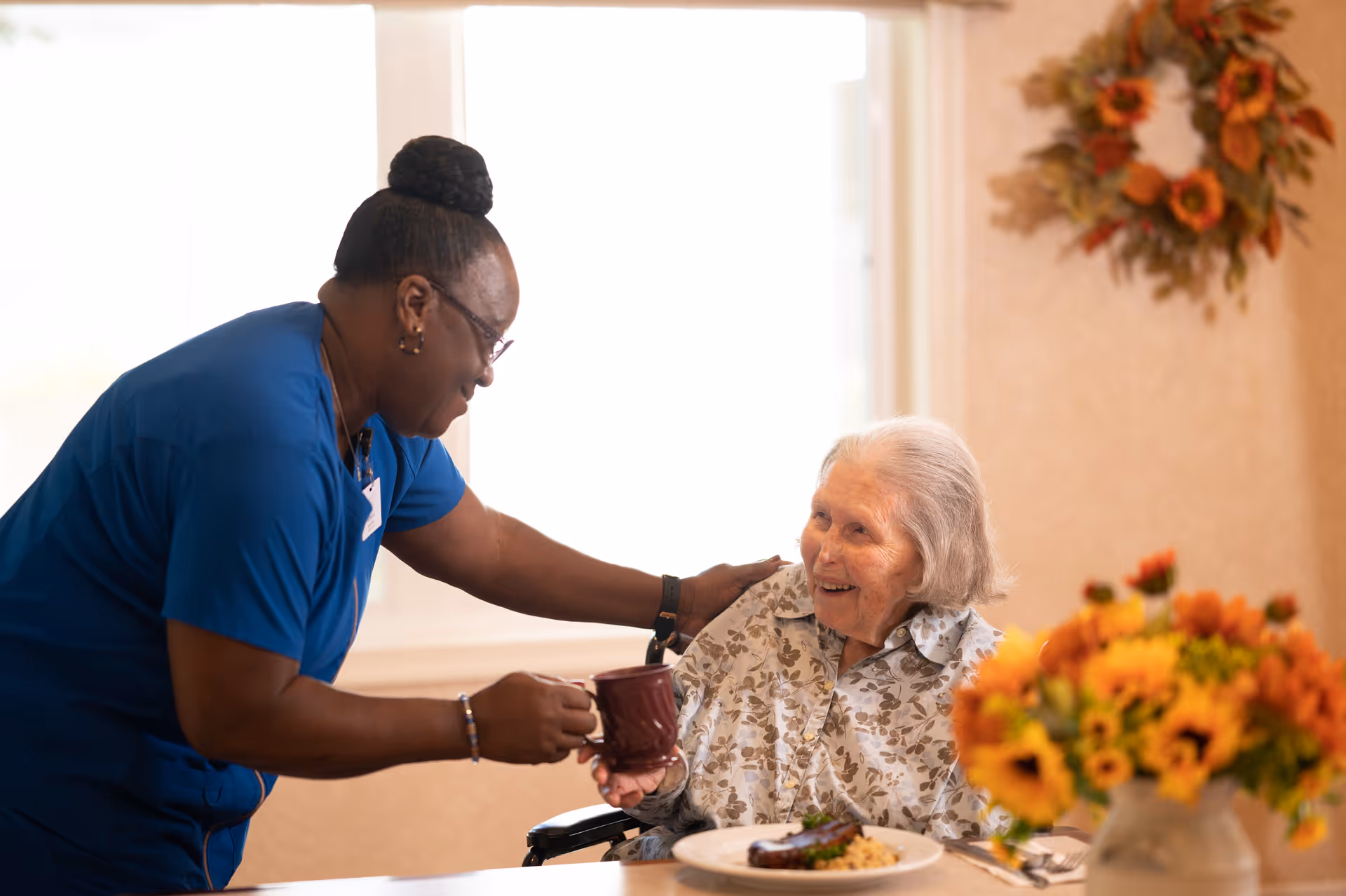 A caregiver in blue scrubs hands a mug to a smiling elderly woman seated at a dining table with a plate of food and a vase of flowers.