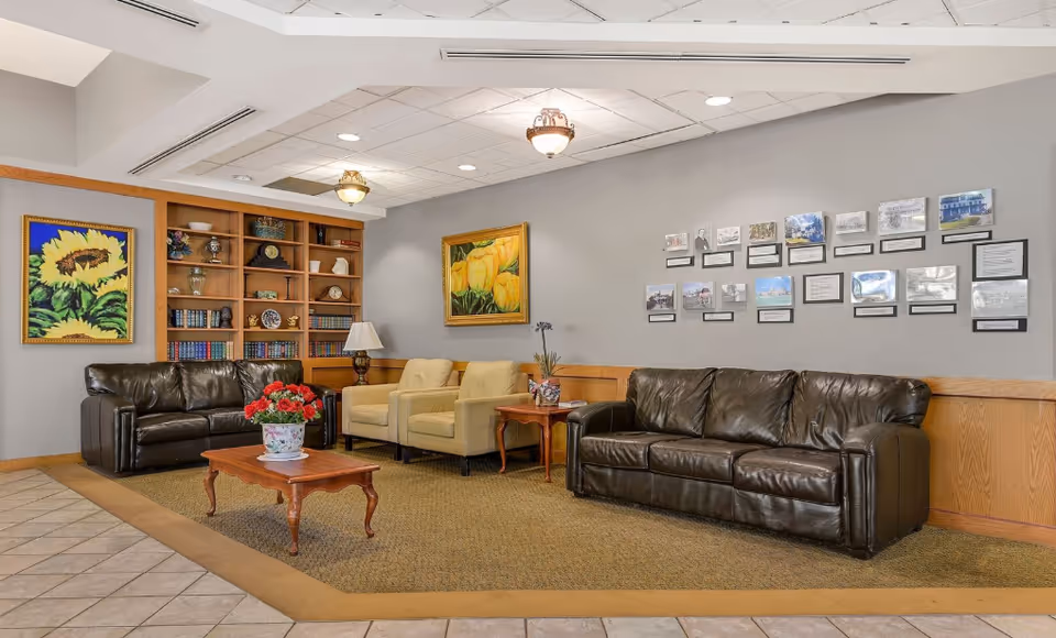 A comfortable lounge area with two dark brown leather sofas, two beige armchairs, a wooden coffee table with a flower pot, and a wooden bookshelf filled with books and decorative items. The walls are decorated with framed paintings of sunflowers and yellow tulips, as well as a series of small framed photos and documents.