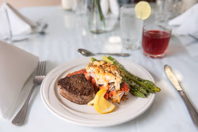A plated meal consisting of a cooked steak, lobster tail with melted cheese on top, asparagus, and a lemon wedge on a white plate. The plate is set on a white tablecloth with a fork, knife, glass of water with a lemon slice, and a glass of red beverage in the background.