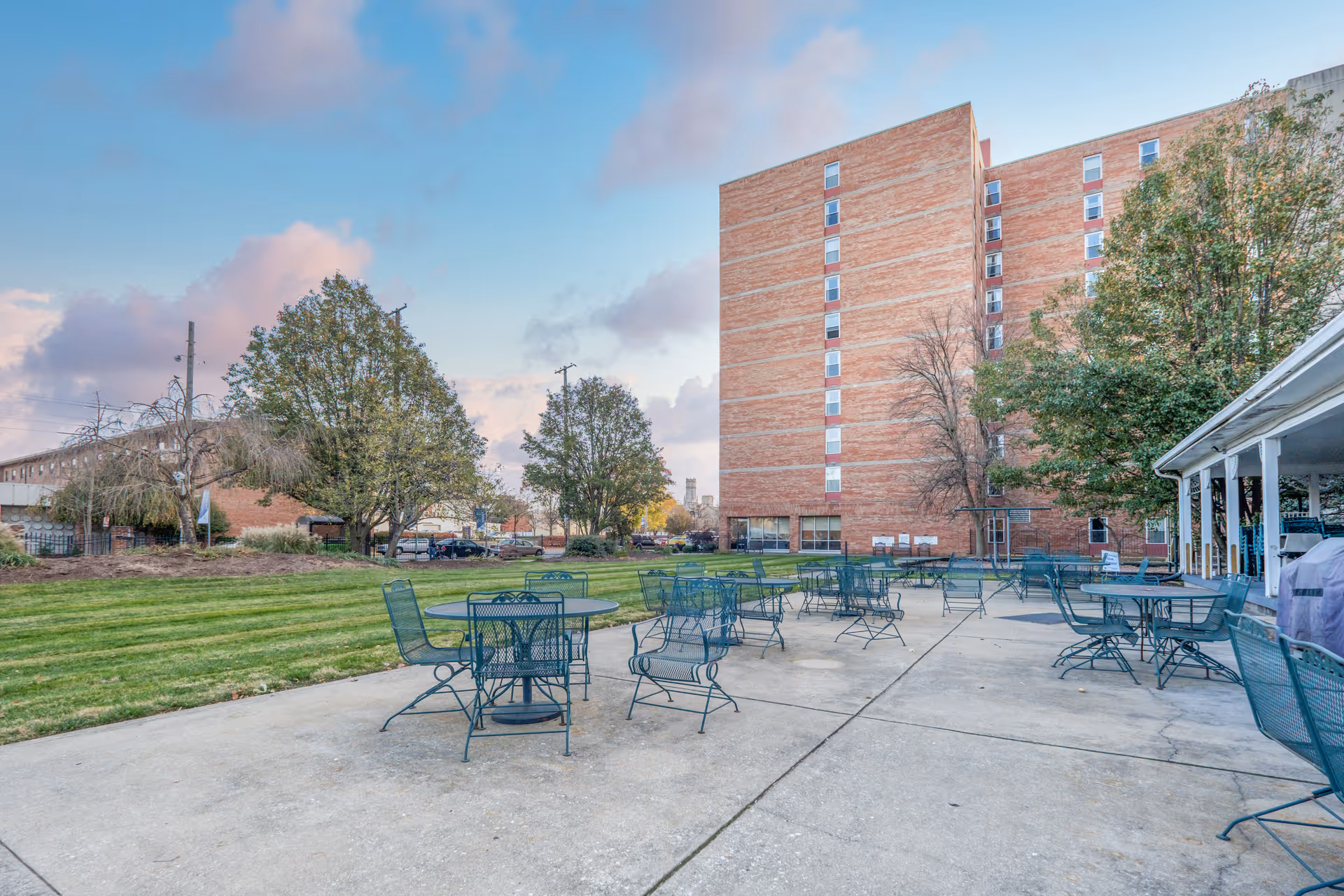 Outdoor patio area with multiple metal tables and chairs on a concrete surface, adjacent to a large brick building. There are trees and a grassy lawn surrounding the patio under a partly cloudy sky.