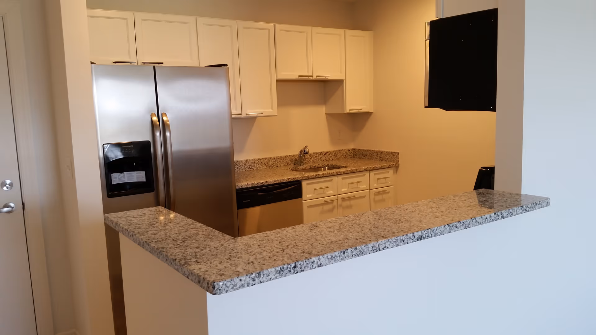 Modern kitchen area with granite countertops, white cabinets, a stainless steel refrigerator with water dispenser, a dishwasher, and a sink. The kitchen has a pass-through counter opening to an adjacent room.