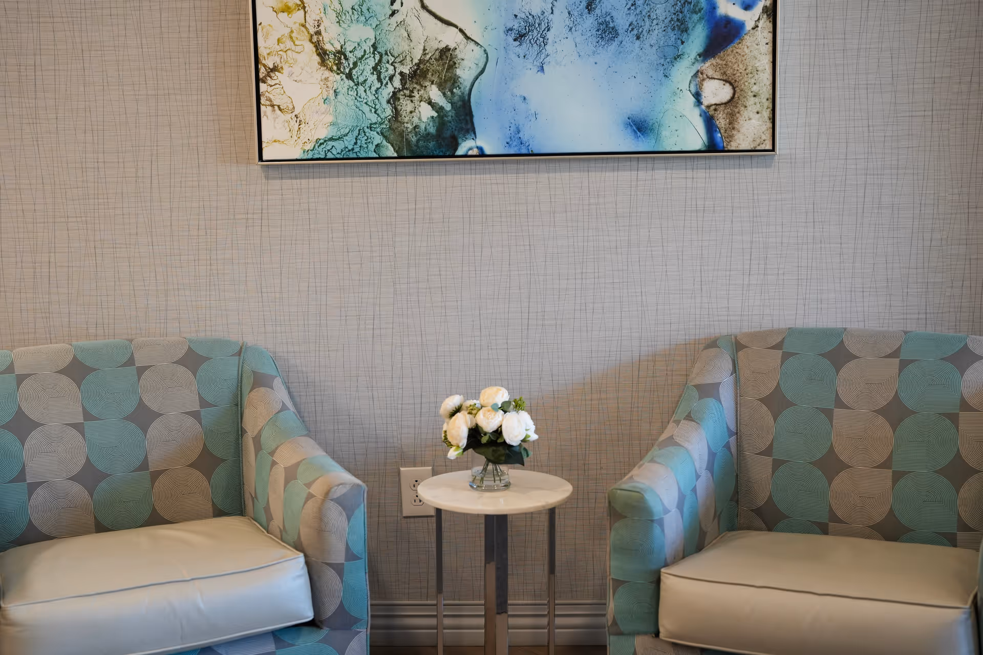 Two patterned armchairs flank a small round table with a vase of white flowers beneath an abstract painting on a textured wall.