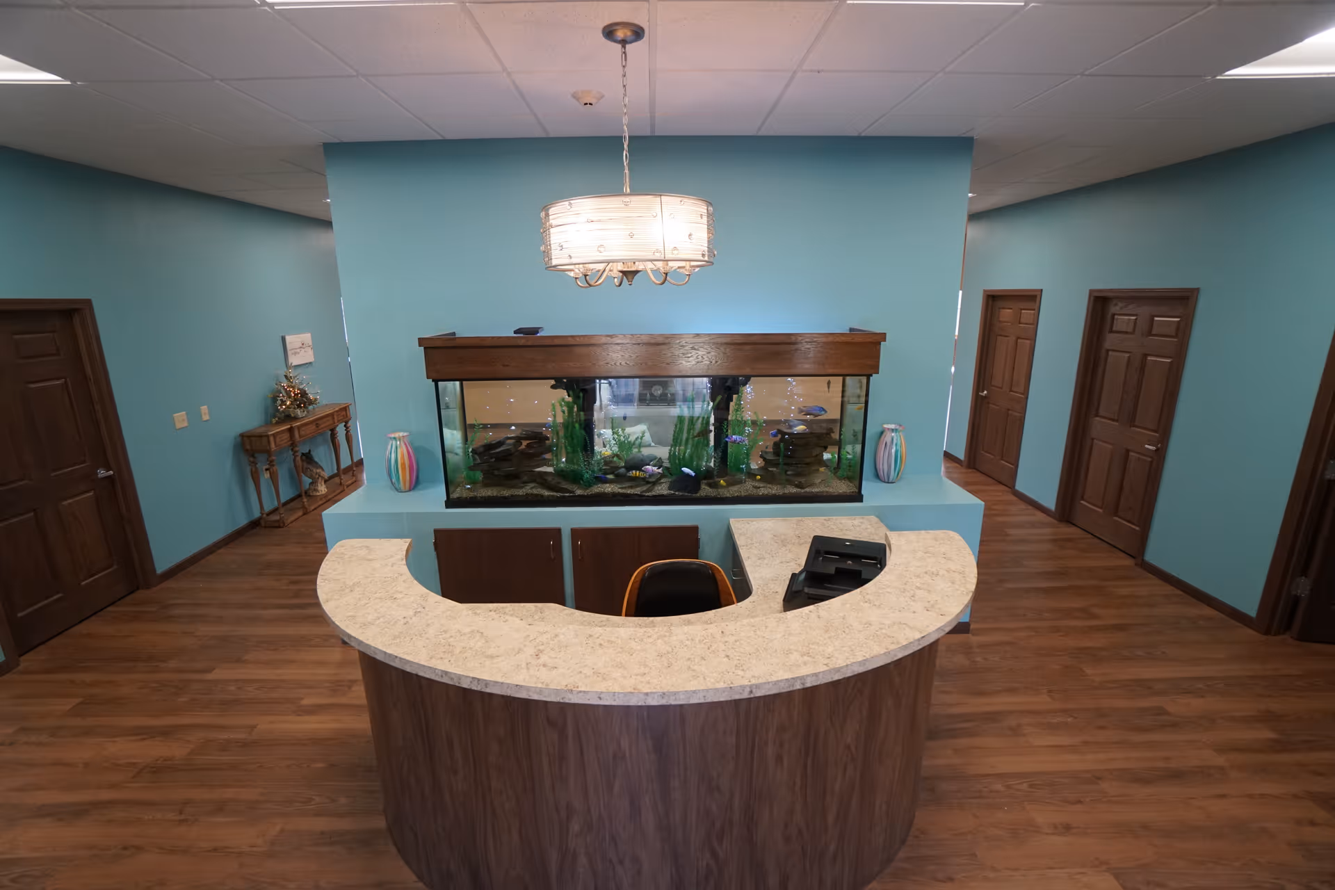 Reception area with a curved wooden desk and a large aquarium built into a blue wall behind it. The room has wooden floors, teal walls, several closed wooden doors, a small table with decorative items on the left side, and a modern hanging light fixture above the aquarium.