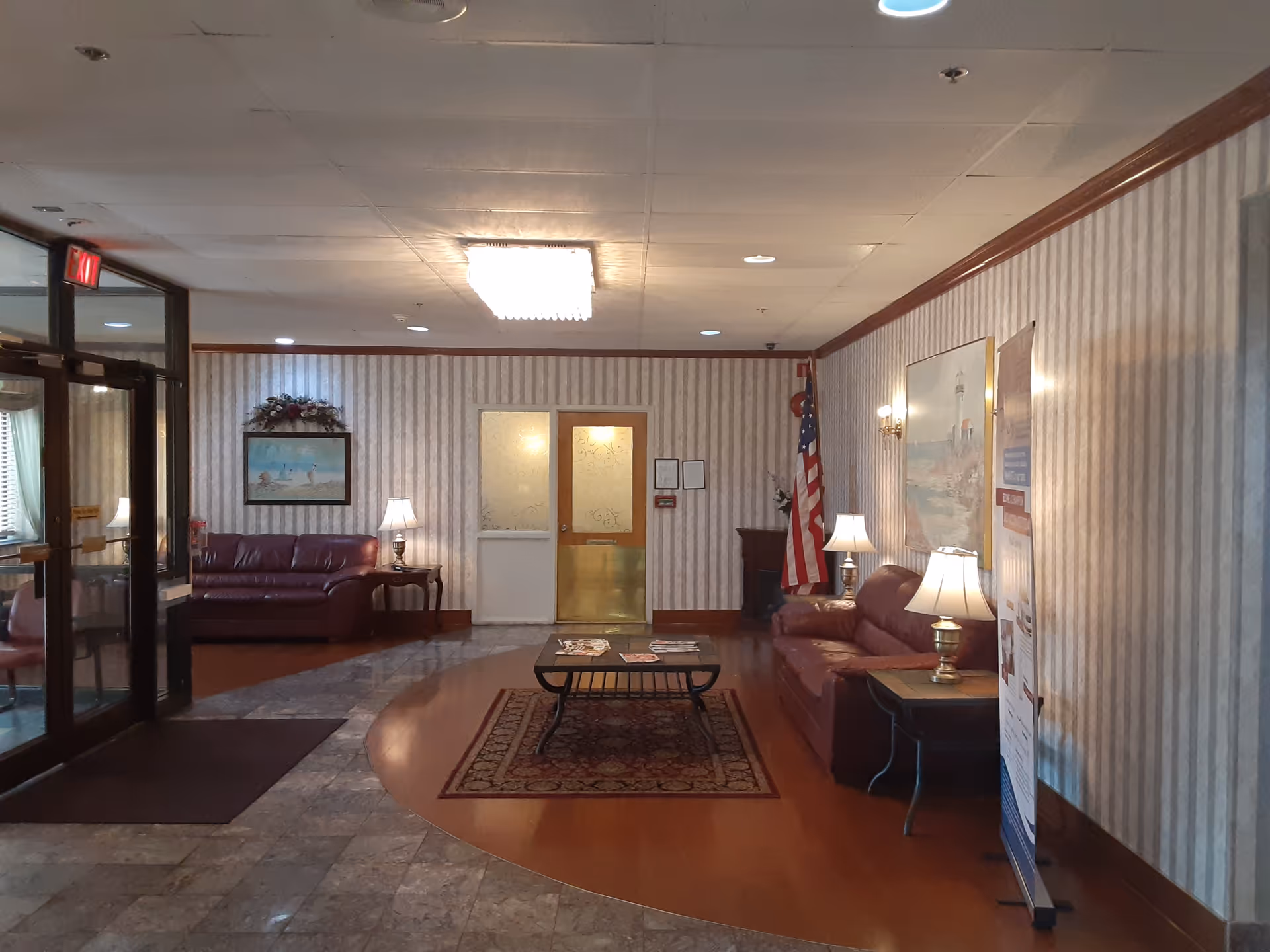 A senior living facility lobby with leather sofas, a coffee table on a rug, lamps, an American flag and a doorway at the far wall.