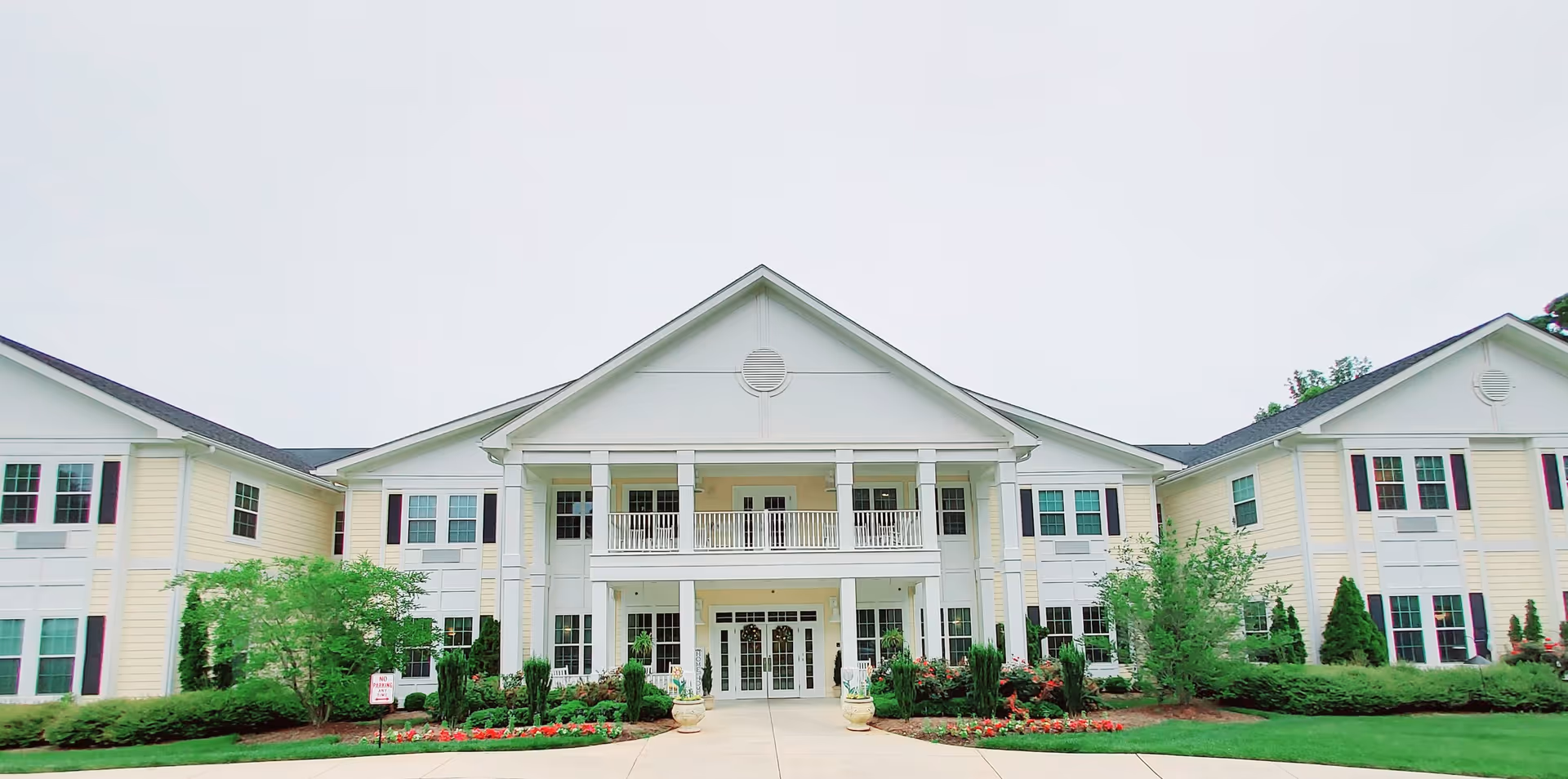 Front exterior view of a large, two-story senior living facility building with white and light yellow siding, multiple windows, a covered entrance with columns, landscaped greenery, and flower beds.