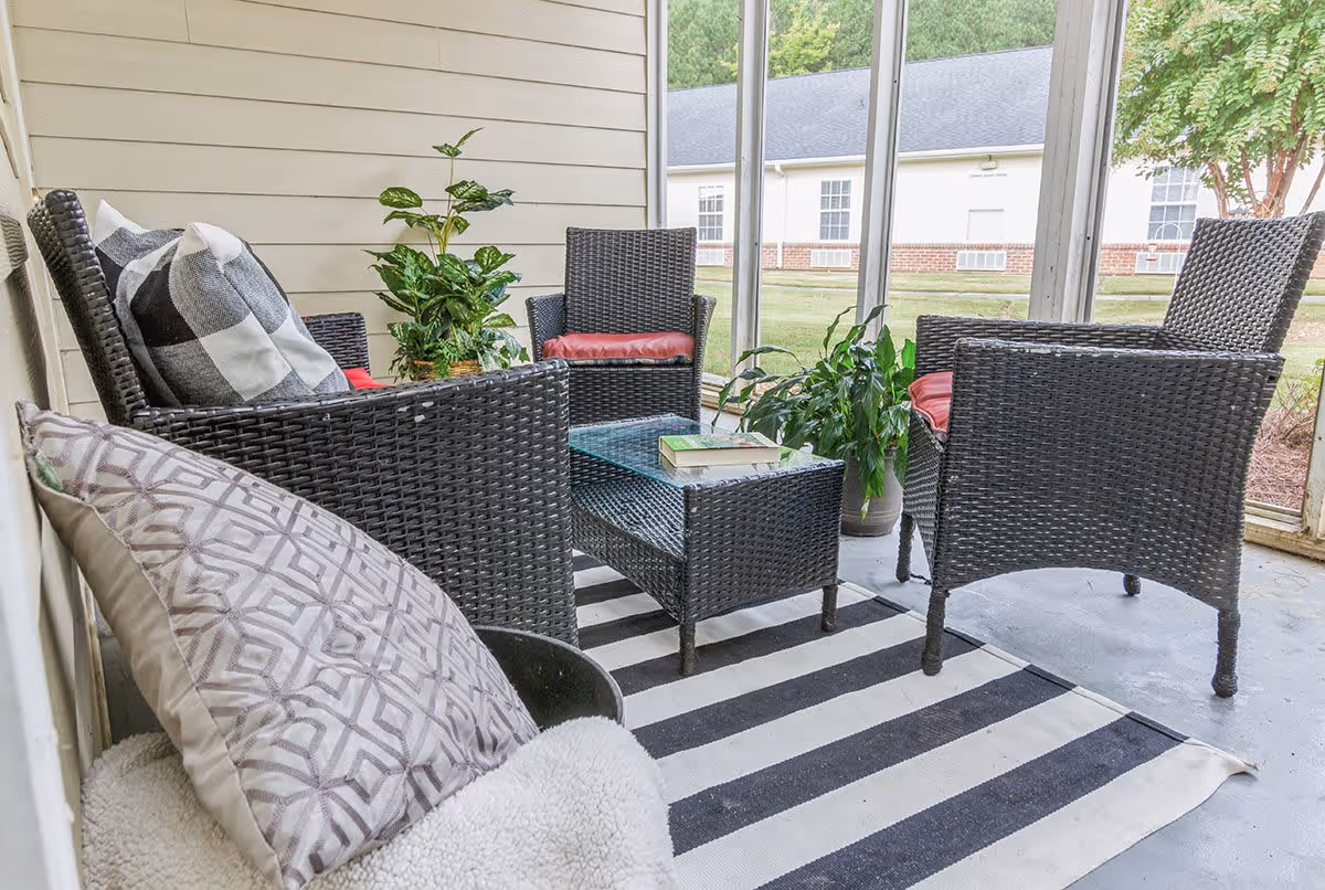 A cozy outdoor seating area with four dark wicker chairs featuring red cushions arranged around a matching wicker coffee table with books on top. The area is decorated with a striped black and white rug and potted plants, enclosed by screened windows overlooking a grassy yard and neighboring buildings.