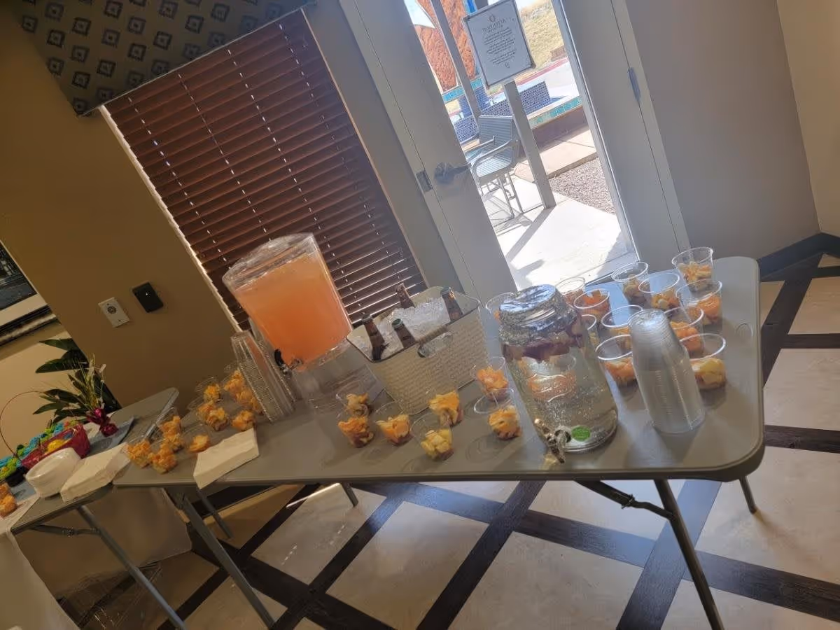 A table set with refreshments including fruit cups, a large drink dispenser with pink lemonade, and a jar of infused water, with a sunny entrance visible in the background.