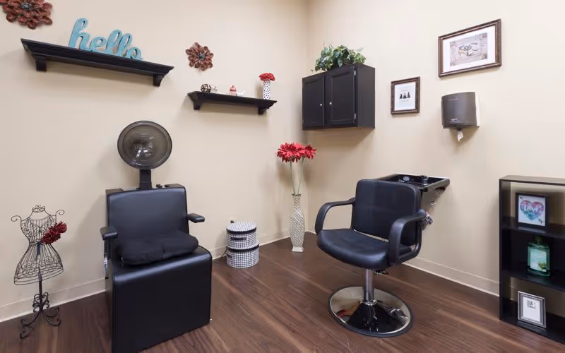 A small salon room with two black salon chairs, one with a hair dryer hood attached. The room has light beige walls with decorative shelves holding small items and flowers. There is a black cabinet mounted on the wall, framed pictures, a paper towel dispenser, and a small black shelving unit with decorative items. The floor is dark wood.