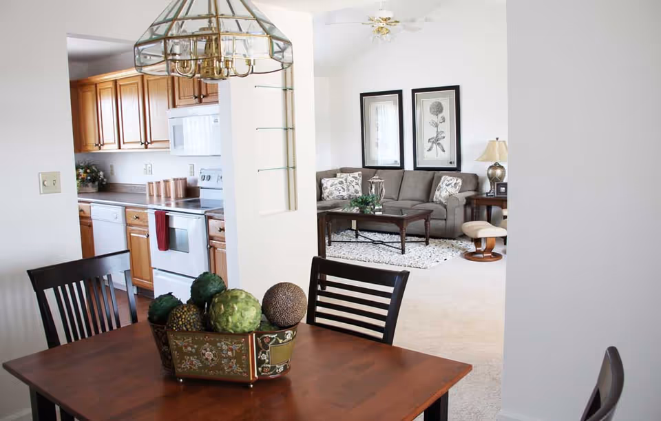Open-plan interior with a wooden dining table and decorative centerpiece in the foreground, a kitchen to the left, and a living room with a sofa in the background.