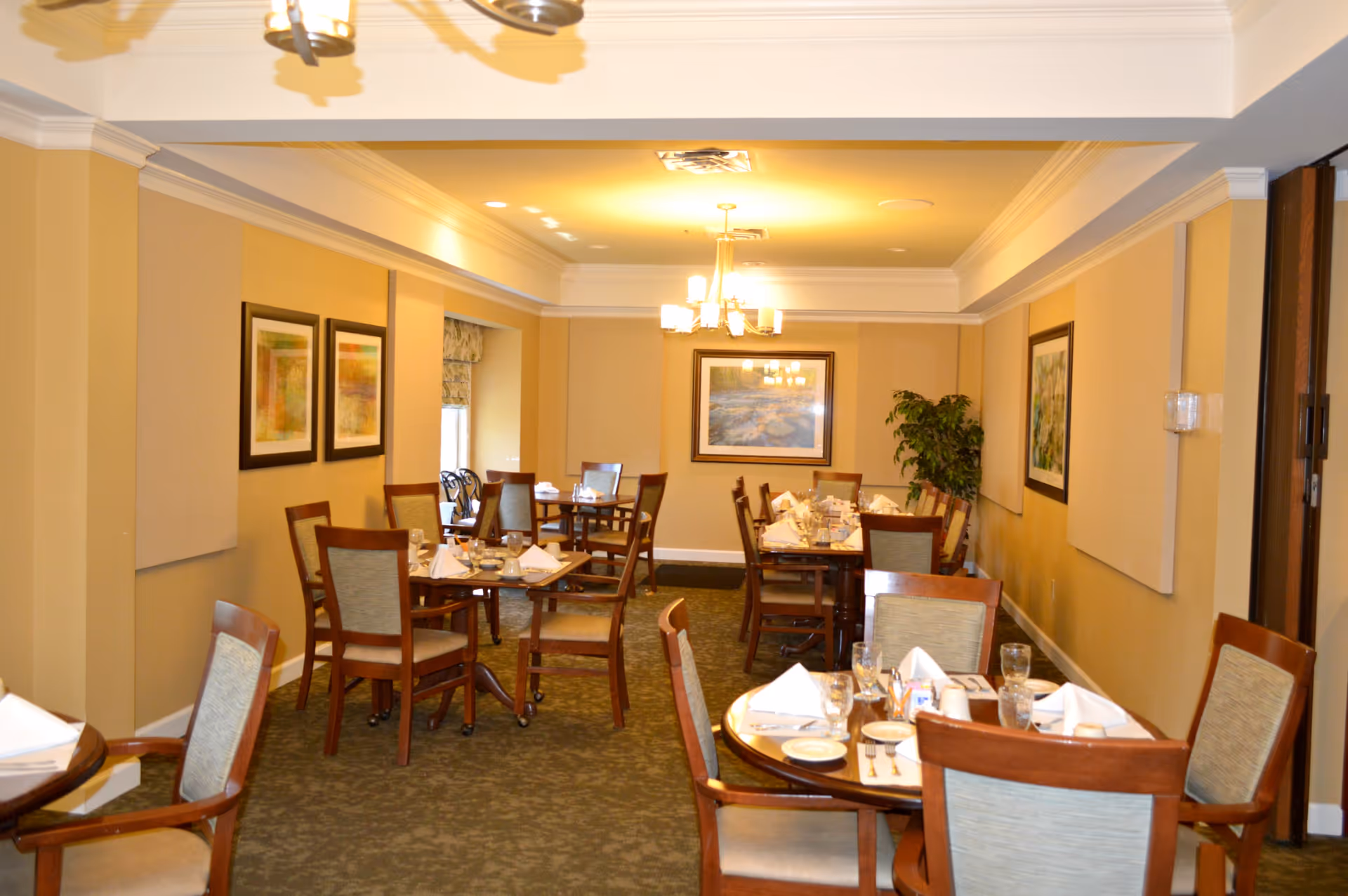 A warmly lit dining room with several wooden tables and chairs arranged neatly. Each table is set with white napkins, glasses, plates, and cutlery. The walls are decorated with framed artwork and there is a potted plant in the corner. The room has a carpeted floor and ceiling lights providing soft illumination.