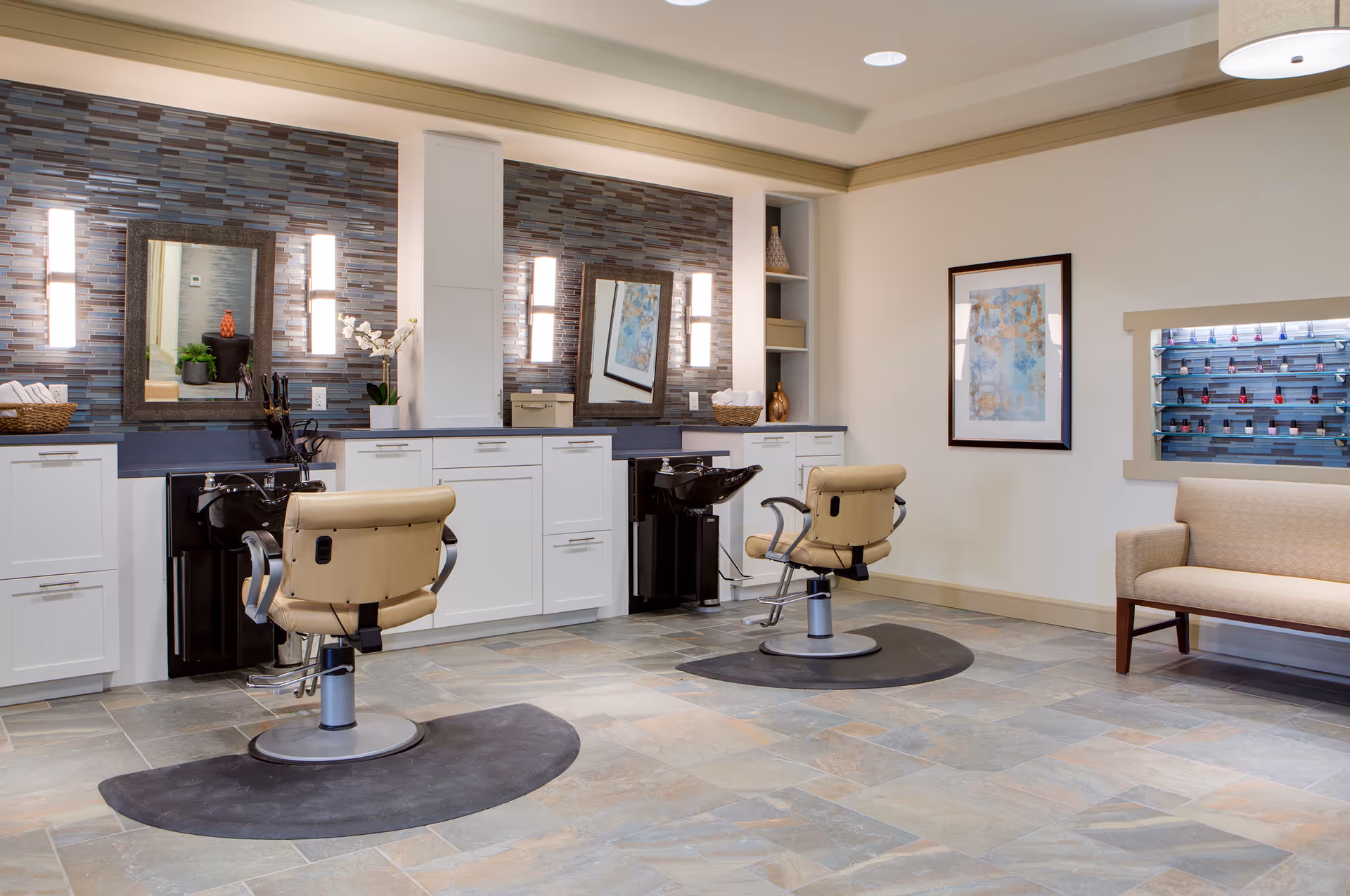 Interior of a salon area in a senior living facility with two beige salon chairs in front of black hair washing sinks. The back wall features a modern tiled backsplash with two mirrors and vertical lights. There are white cabinets below the sinks and a beige cushioned bench on the right side. A wall-mounted rack holds various nail polish bottles.