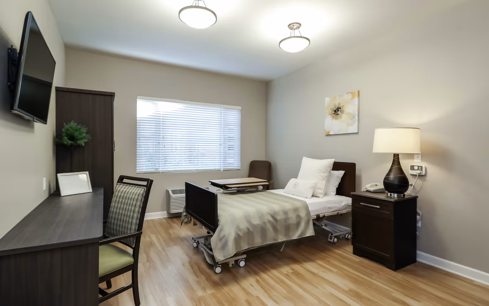A clean and well-lit nursing home bedroom with a single hospital-style bed, a bedside table with a lamp and telephone, a chair, a desk with a chair, a wall-mounted TV, and a window with blinds. The room has light-colored walls and wooden flooring.