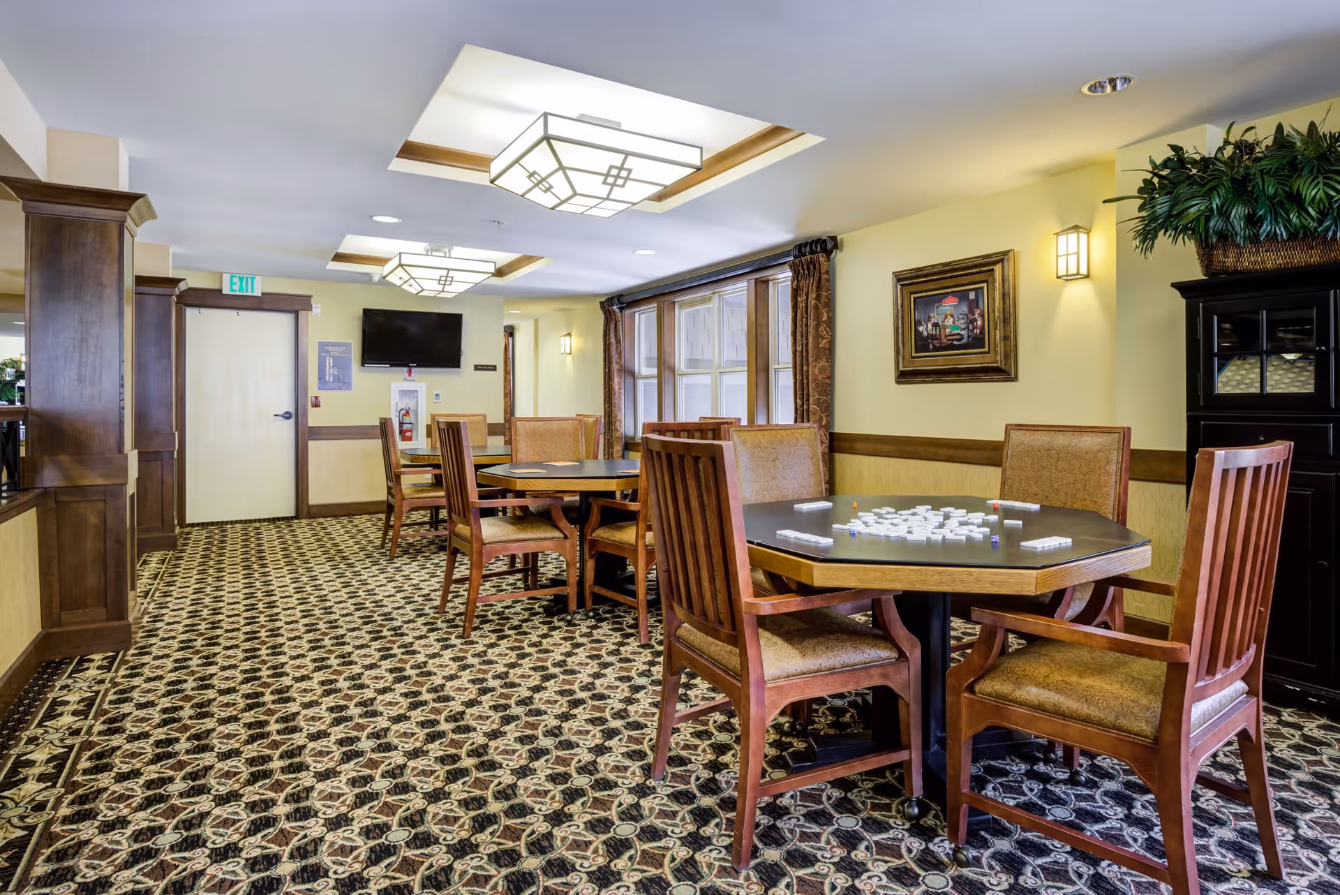 Interior view of a common area in a senior living facility with multiple wooden tables and chairs arranged on a patterned carpet. One table has a game set up with tiles. The room has yellow walls, framed artwork, large windows with curtains, ceiling lights, and a wall-mounted TV near an exit door.
