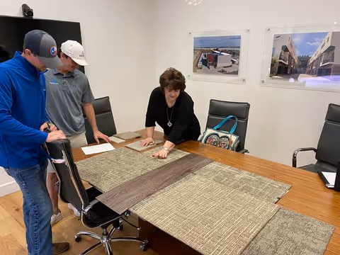 Three people in a meeting room examining different flooring samples laid out on a wooden conference table. The room has black office chairs, white walls, and two framed pictures of buildings on the wall.