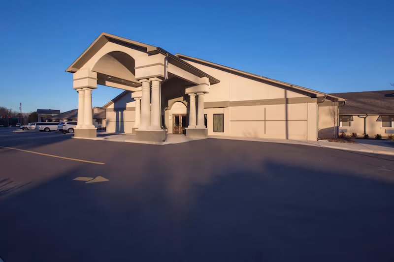 Front entrance of a beige nursing and rehab center with a covered porte-cochère and empty parking lot under a clear blue sky.