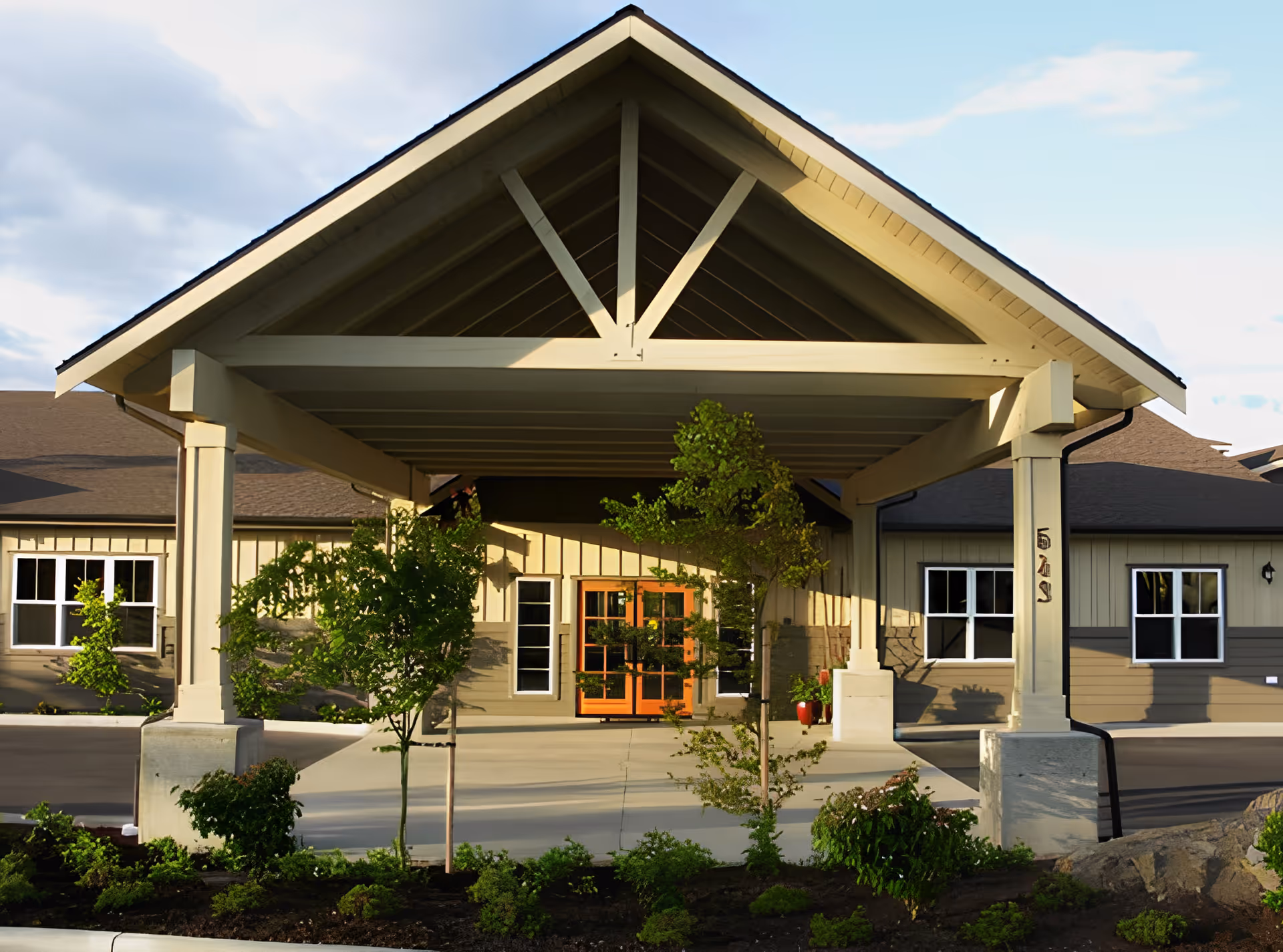 Entrance of a senior living facility with a large covered driveway supported by white pillars, surrounded by small trees and shrubs, with a building featuring beige siding and multiple windows in the background.