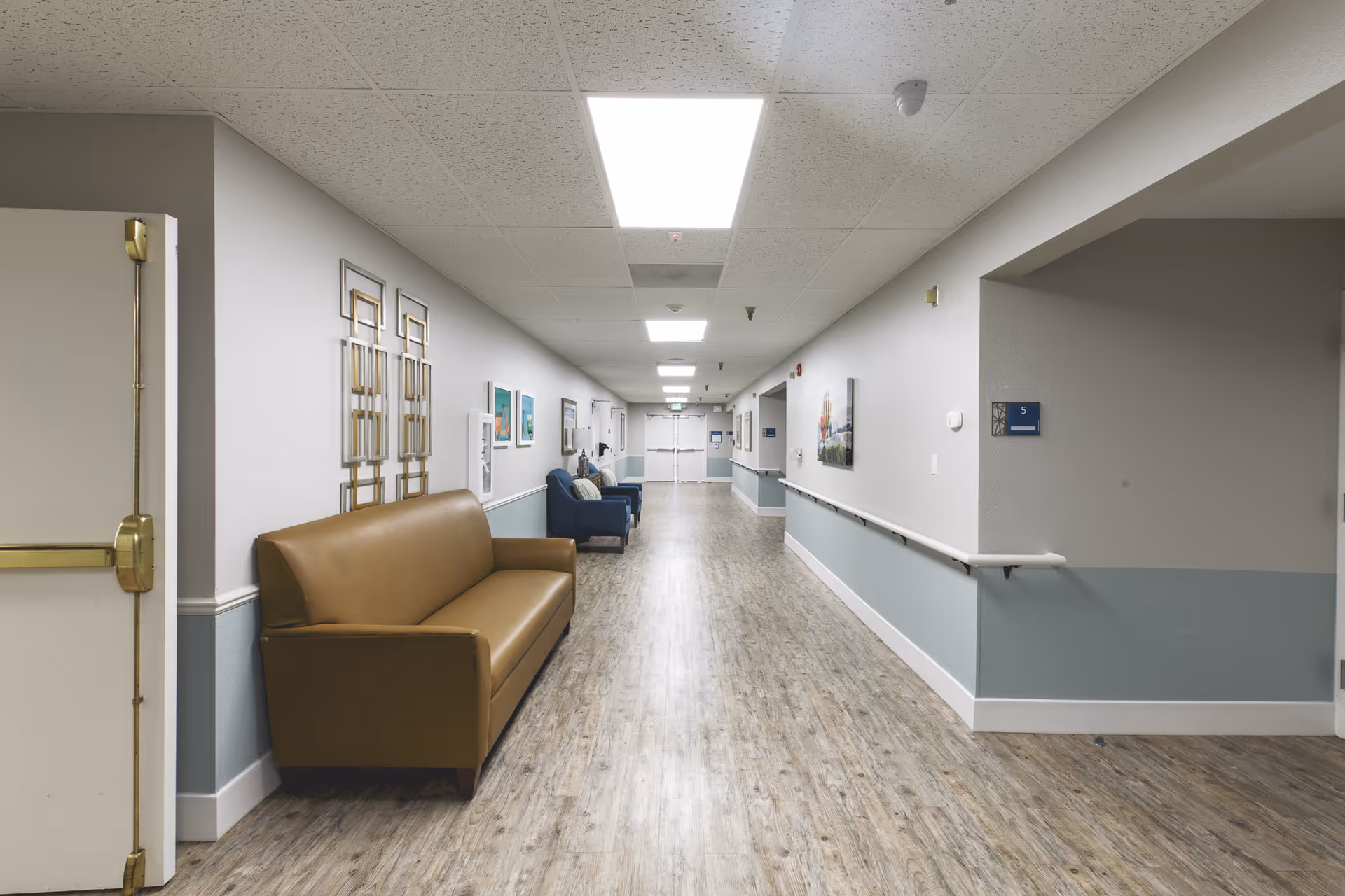 A long, well-lit hallway in a senior living facility with light gray and blue walls, wood-patterned flooring, and ceiling lights. There are brown and blue couches along the left side, decorative wall art, and handrails on the right wall. Double doors are visible at the far end of the hallway.