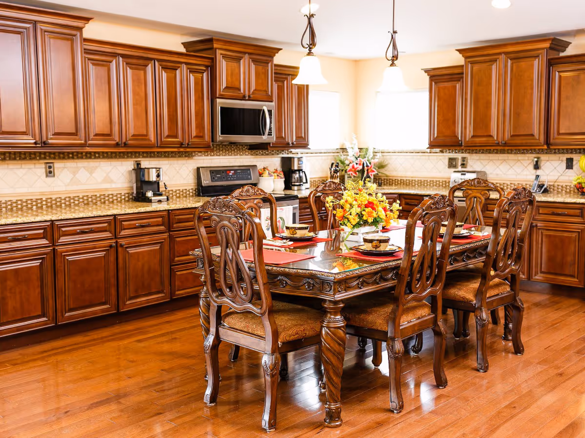 A spacious kitchen with polished wooden cabinets and hardwood floors. In the center, there is an ornate wooden dining table set with six matching chairs, each with intricate carvings. The table is decorated with a floral centerpiece and place settings including plates, bowls, and cups. The kitchen features modern appliances including a microwave, stove, coffee maker, and toaster. Two pendant lights hang from the ceiling, illuminating the area.