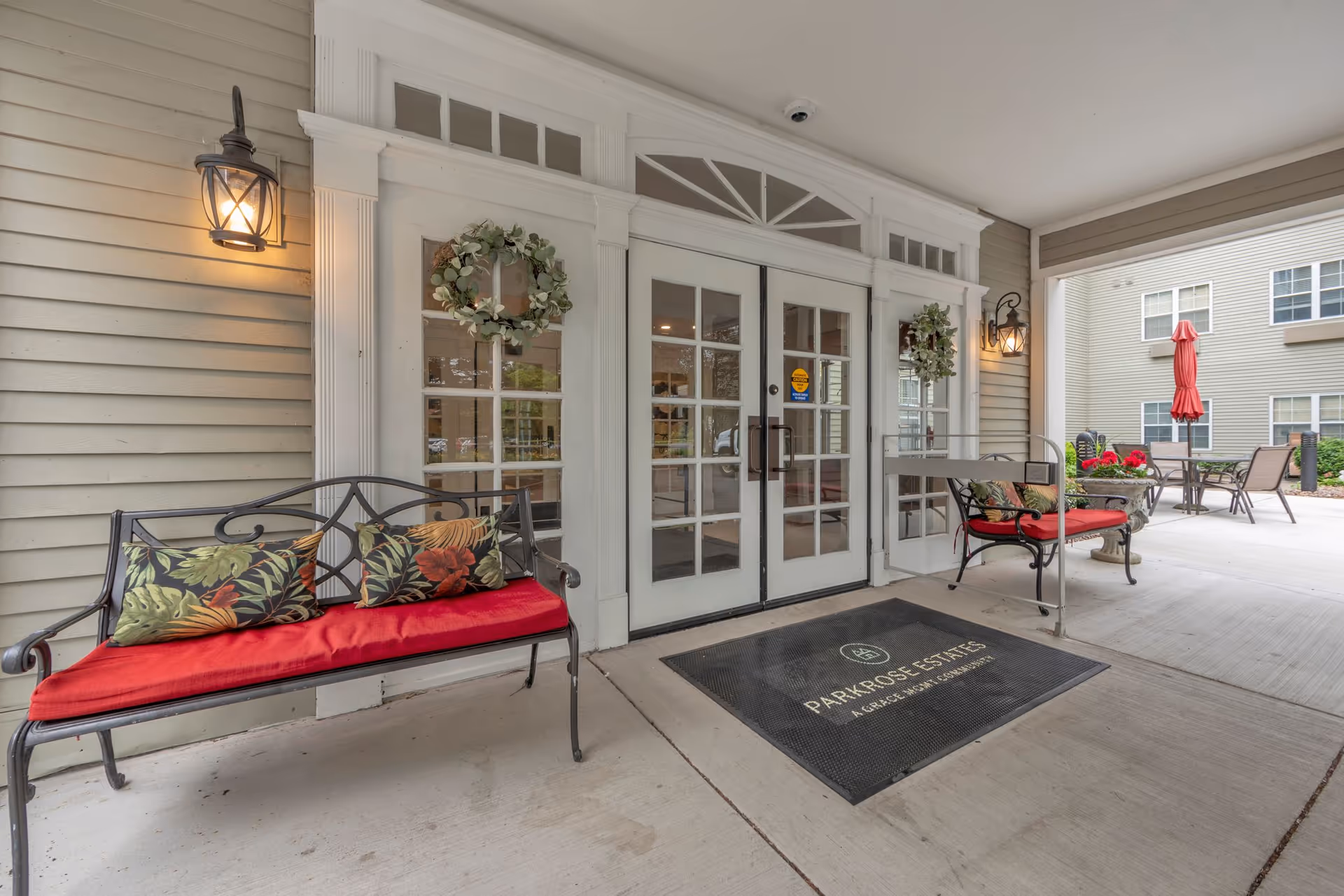 Covered entrance area of Parkrose Estates featuring double glass doors with wreaths, two black metal benches with red cushions and floral pillows, wall-mounted lantern lights, and outdoor seating with tables and chairs in the background.