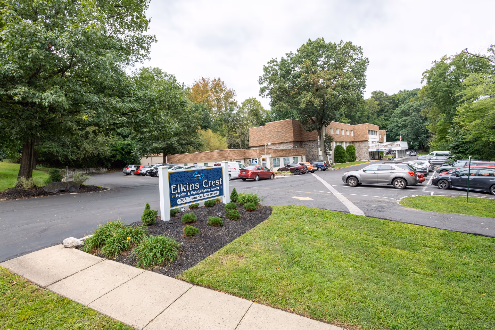 Exterior view of Elkins Crest Health & Rehabilitation Center showing the building, parking lot with several cars, a sidewalk, green grass, trees, and a sign with the facility's name and address.