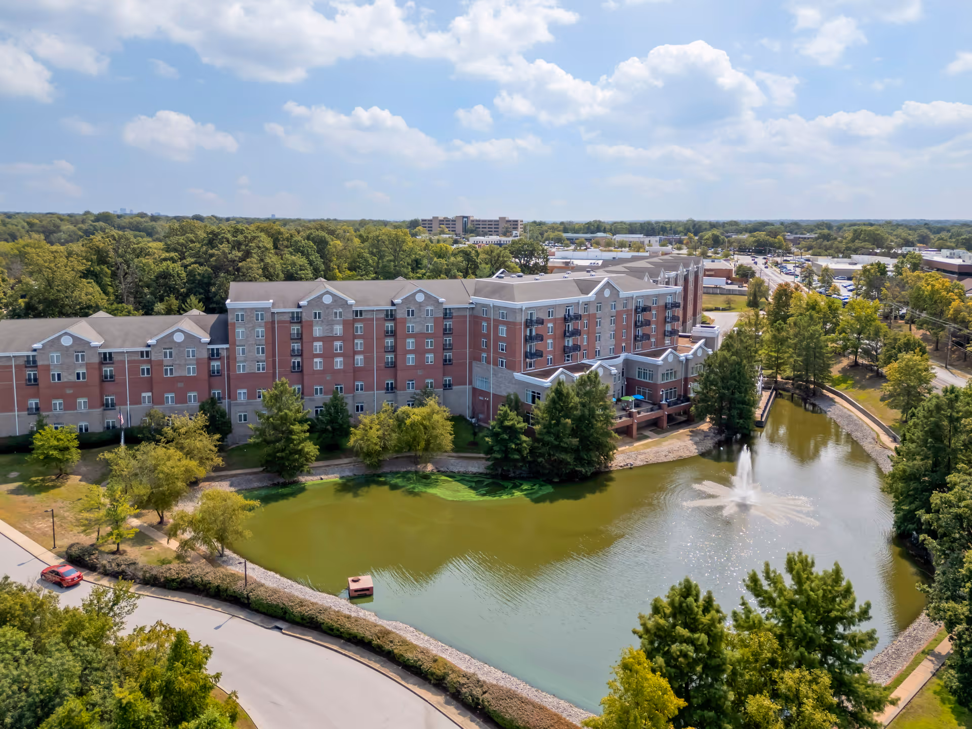 Aerial view of a large multi-story senior living facility building named Brookdale Creve Coeur, situated next to a pond with a water fountain. The building is surrounded by trees and greenery, with a road and parking area visible nearby under a partly cloudy sky.