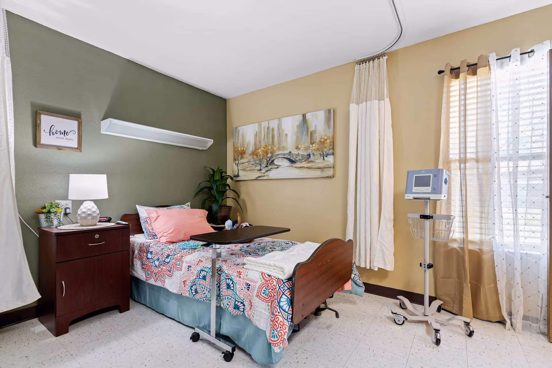 A nursing facility bedroom with a single bed featuring colorful bedding and pink pillows. Next to the bed is a wooden nightstand with a lamp and a small plant. A medical monitor on a stand is positioned near a window with sheer and beige curtains. The walls are painted green and beige, decorated with a framed picture and a painting of a cityscape with a bridge and trees.