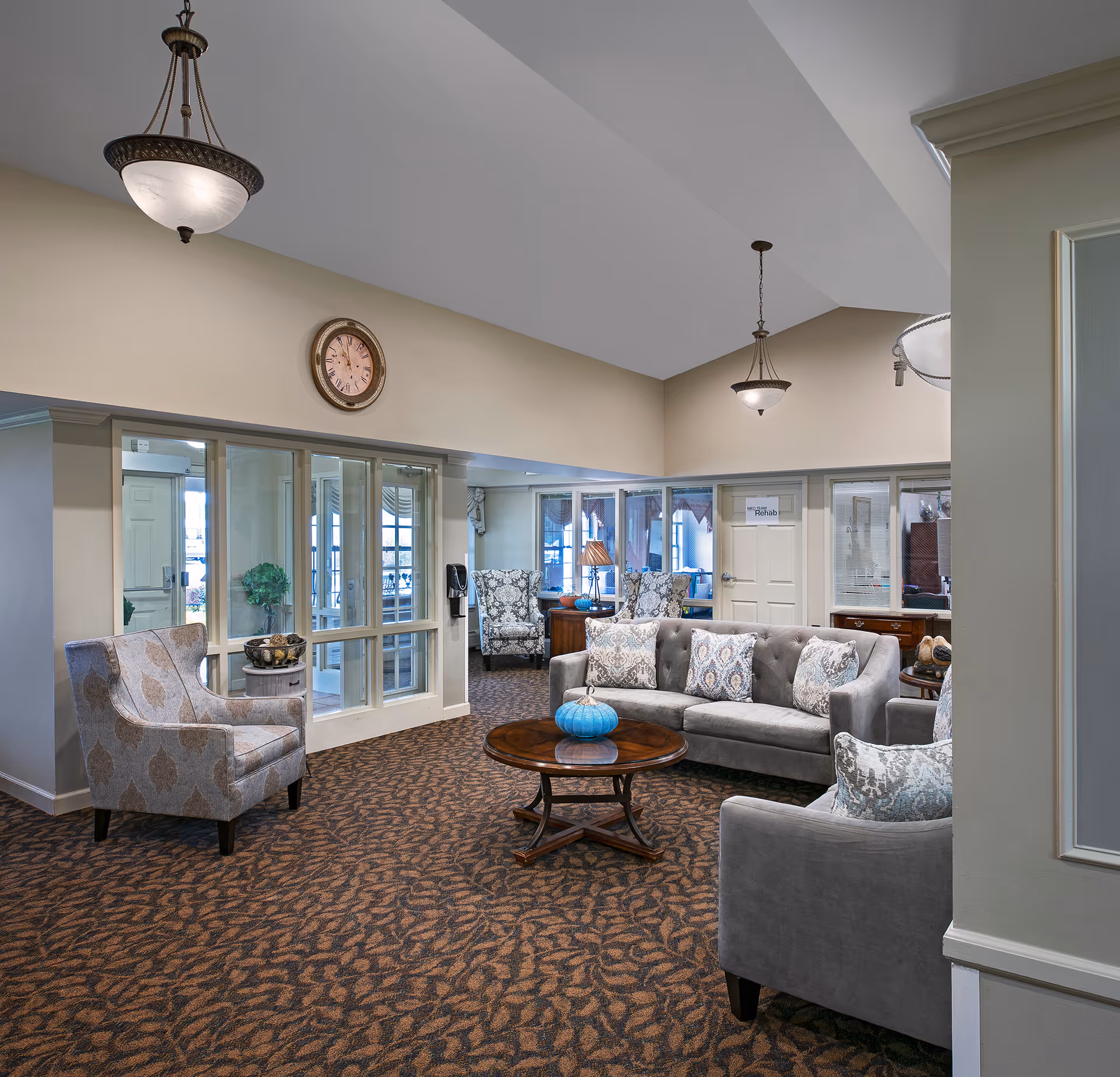 Bright communal lounge with upholstered sofas and chairs arranged around a wooden coffee table under hanging lights.