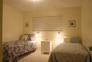 Softly lit bedroom with two twin beds separated by a nightstand, wall-mounted lamps, and a window with blinds.