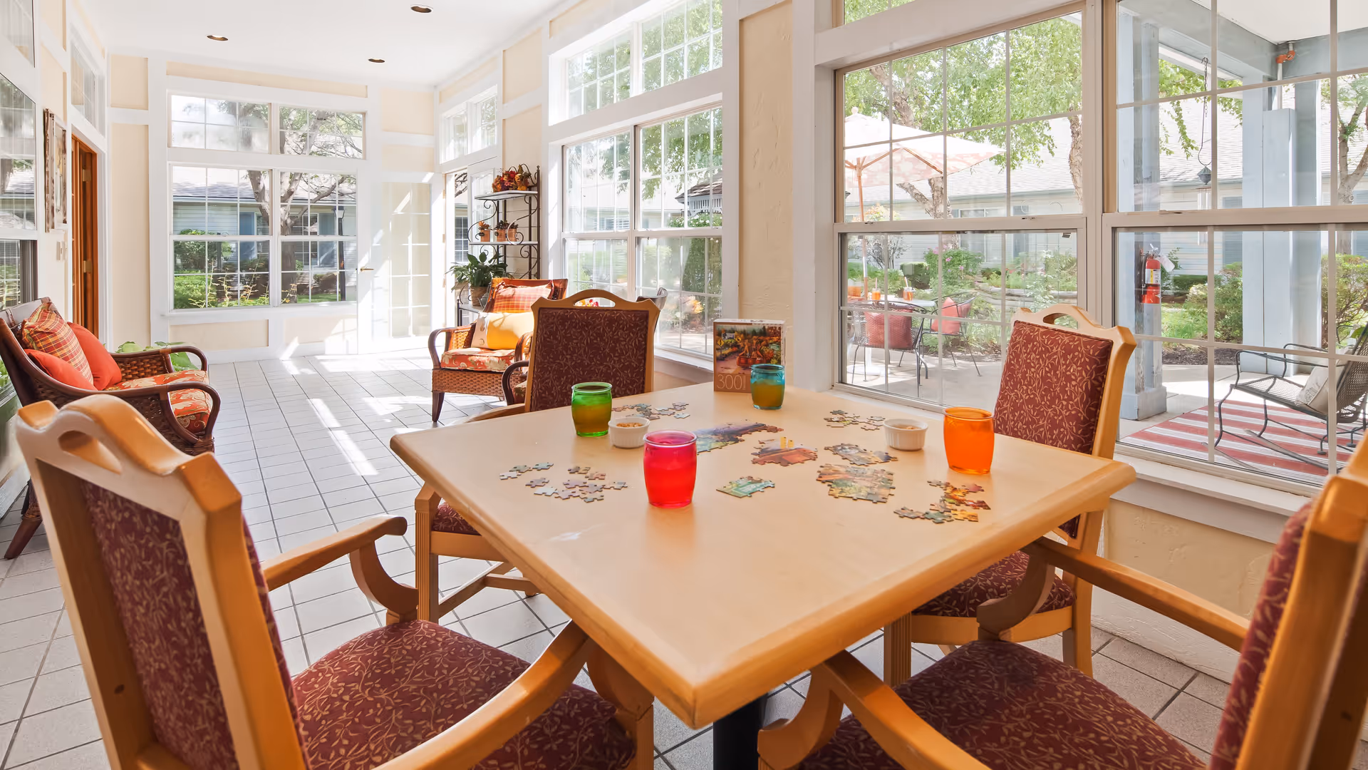 A bright and sunny common area with large windows overlooking a garden patio. The room features a wooden table with four chairs around it, each chair having a patterned cushion. On the table, there is a partially completed jigsaw puzzle and four colorful drinking glasses. In the background, there are additional chairs with cushions and a metal shelving unit with decorative items.