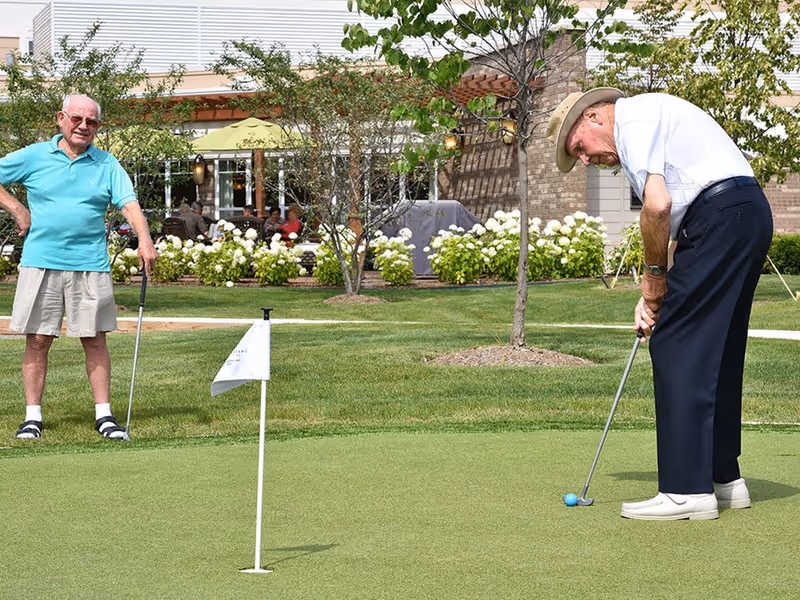 Two elderly men playing golf on a putting green outside a senior living facility. One man is putting while the other watches, with a building, trees, and flowering bushes in the background.