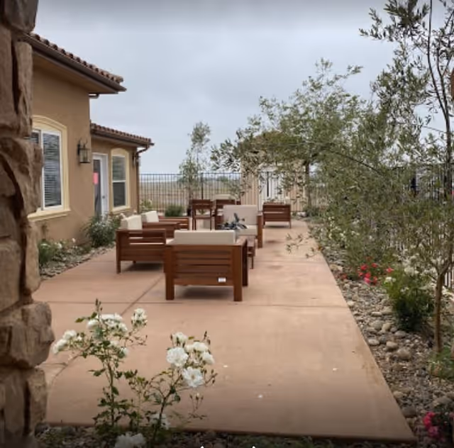 Outdoor patio area with wooden cushioned chairs and tables arranged on a concrete surface next to a building. There are plants and small trees along the edge of the patio, with a fence in the background under a cloudy sky.