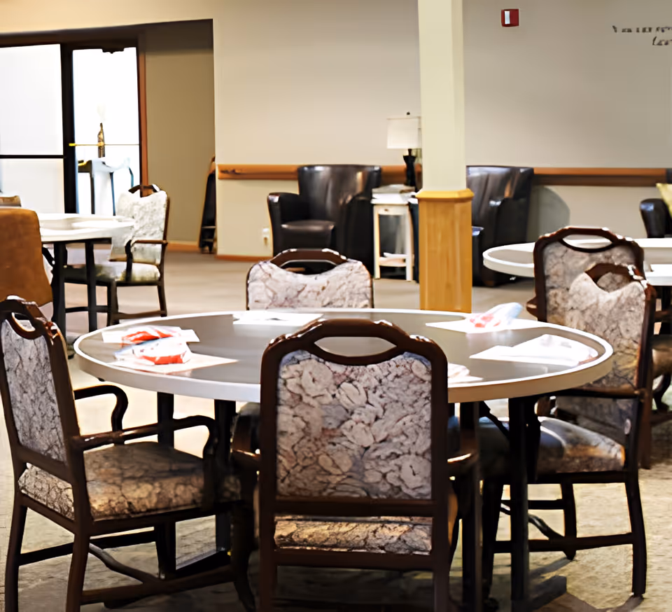 Interior view of a senior living facility dining area with round tables and cushioned chairs. The room has neutral-colored walls, a carpeted floor, and some armchairs and side tables in the background. There is a door with glass panels letting in natural light.