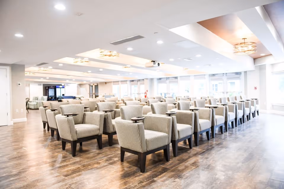 A spacious, well-lit room with rows of beige upholstered chairs, each equipped with a small attached table, arranged neatly on a wooden floor. The ceiling features recessed lighting and decorative light fixtures, and large windows along one side allow natural light to fill the space.