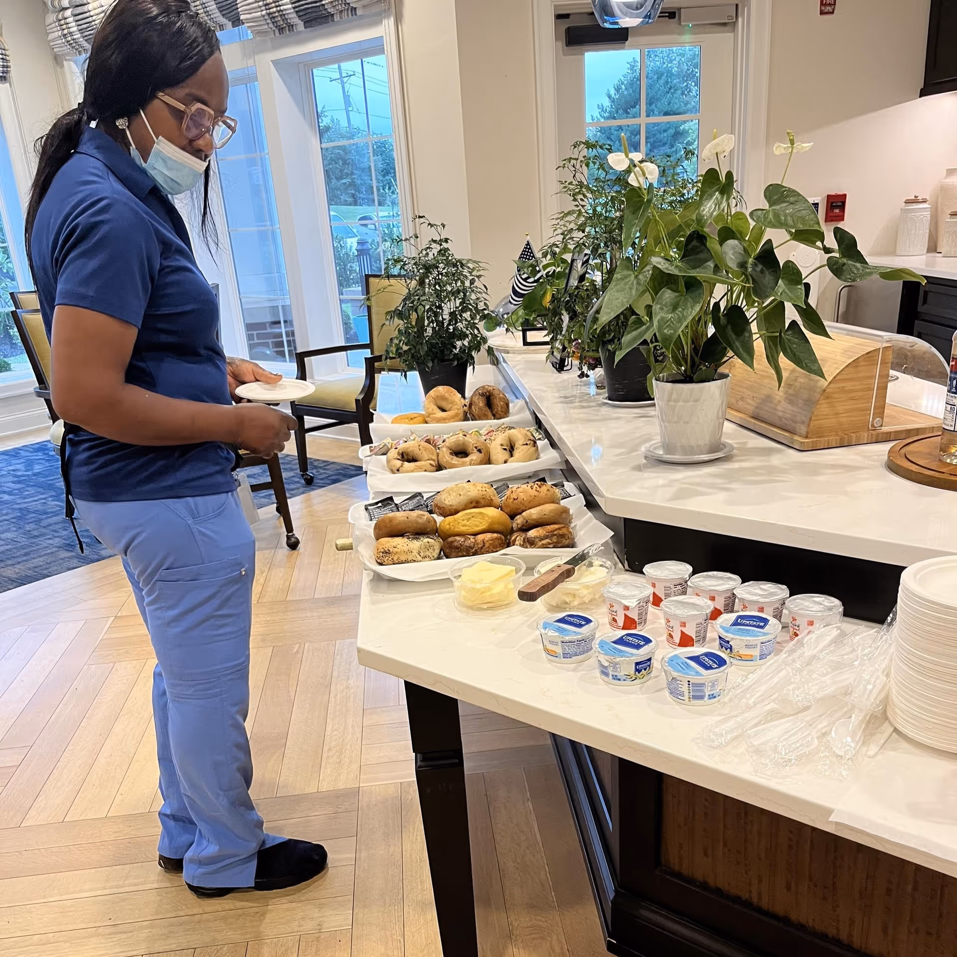 A woman wearing a blue uniform and a face mask stands near a countertop with an assortment of bagels, cream cheese containers, butter, and plastic utensils in a well-lit room with large windows and plants on the counter.