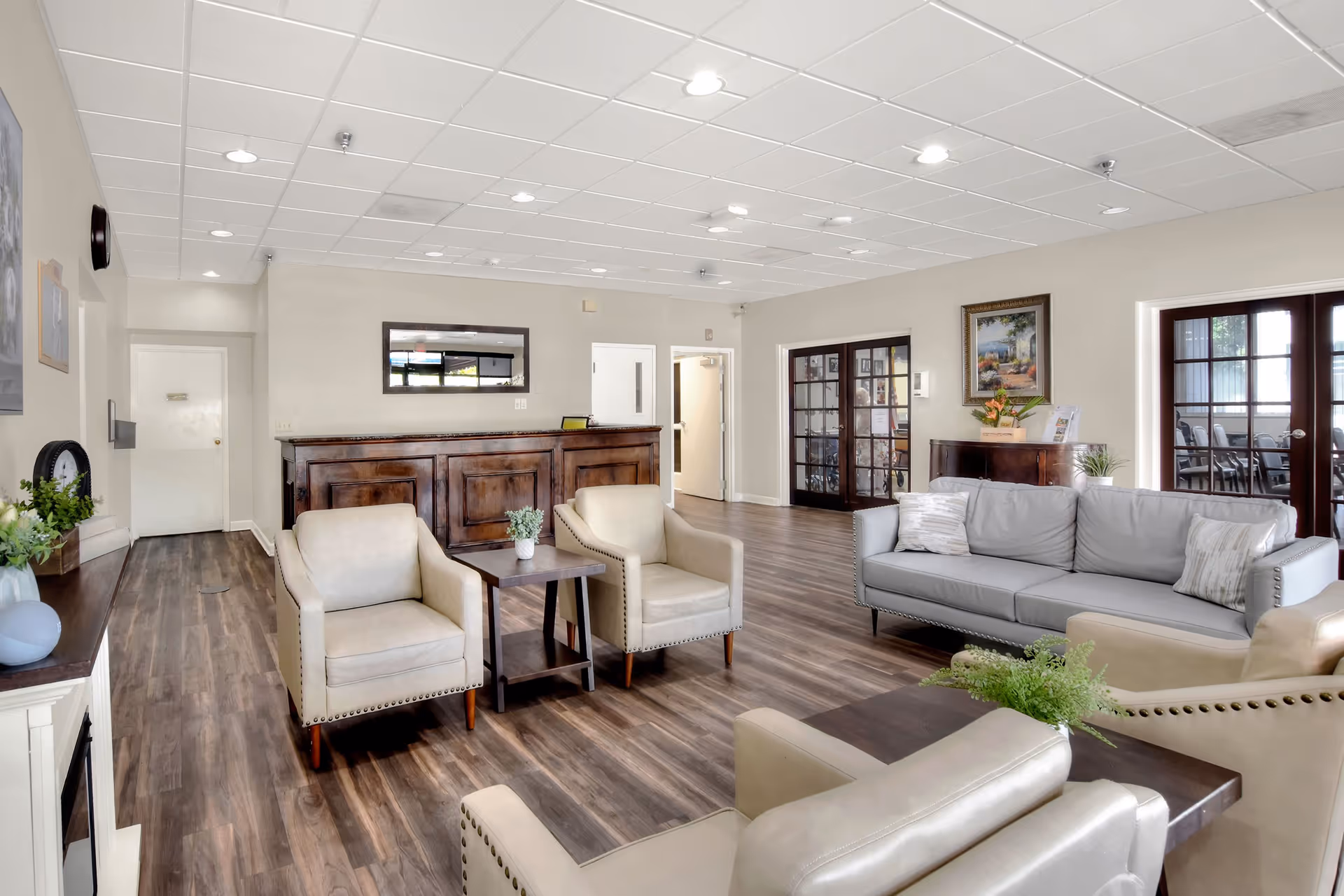A spacious and well-lit common area with wooden flooring, featuring a gray sofa, three beige armchairs, and two dark wooden side tables with small plants. There is a wooden reception desk against the far wall with a rectangular mirror above it. The room has white walls, a white tiled ceiling with recessed lighting, and two sets of glass double doors on the right side. A framed painting and a small cabinet with flowers and brochures are also visible.
