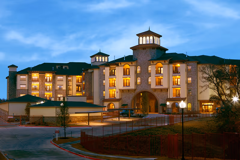 Exterior view of a large, multi-story senior living facility building at dusk with lights glowing warmly from the windows. The building features stone and stucco walls, arched entryways, and decorative towers. There is a driveway and parking area in front, with some trees and fencing visible.