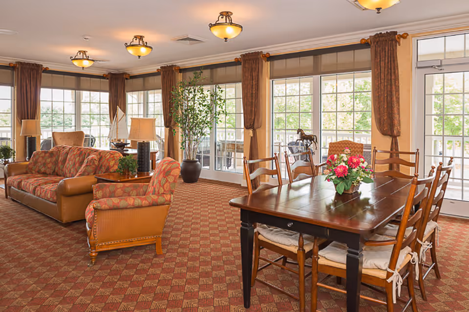 A bright and spacious common area in a retirement village featuring a wooden dining table with six chairs, a floral centerpiece, a patterned carpet, a sofa and armchair with leaf-patterned upholstery, several table lamps, large windows with brown curtains, and a potted plant near the windows.