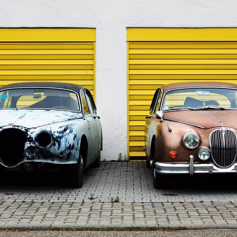Two vintage cars parked side by side in front of bright yellow garage doors on a paved lot.