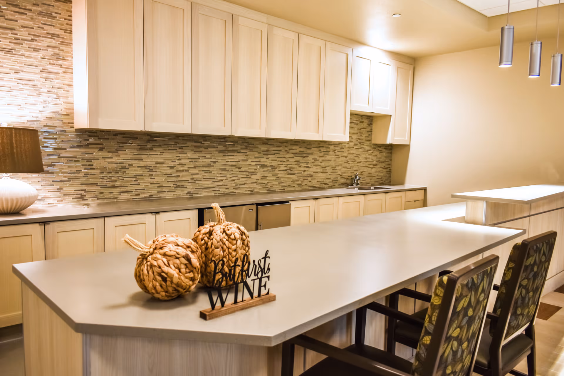 Modern communal kitchen and bar area with a long island, decorative woven pumpkins, and upholstered bar chairs.