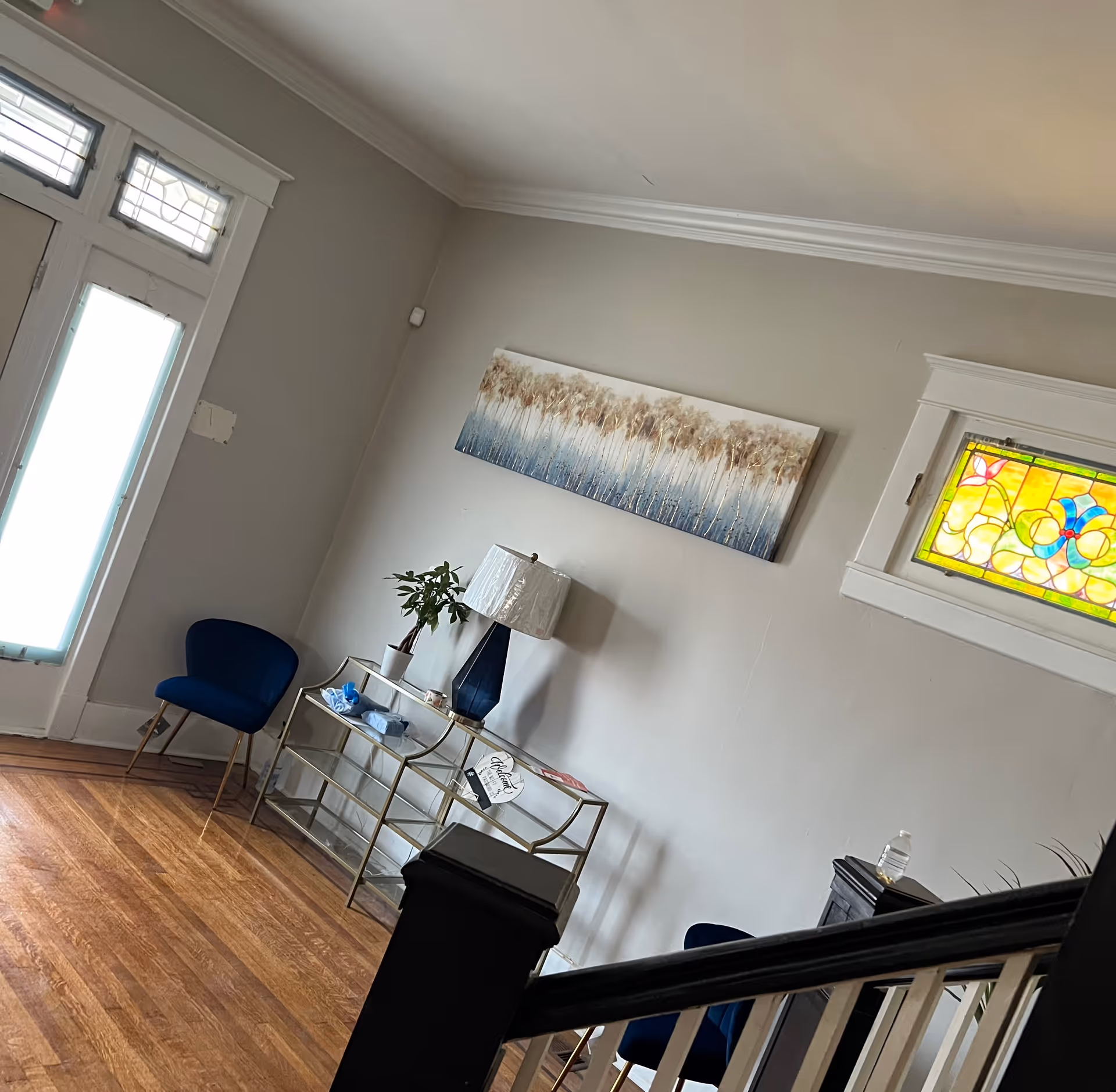 Interior view of a living room area with wooden flooring, a glass console table holding a blue lamp, a small plant, and some items. Two blue chairs are placed near the walls. A colorful stained glass window and a rectangular painting with a nature theme hang on the light gray walls. A staircase railing is visible in the foreground.