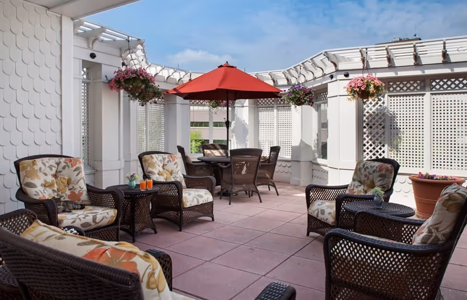 Sunny outdoor patio with wicker chairs and floral cushions, a central table under a red umbrella, and hanging flower baskets.